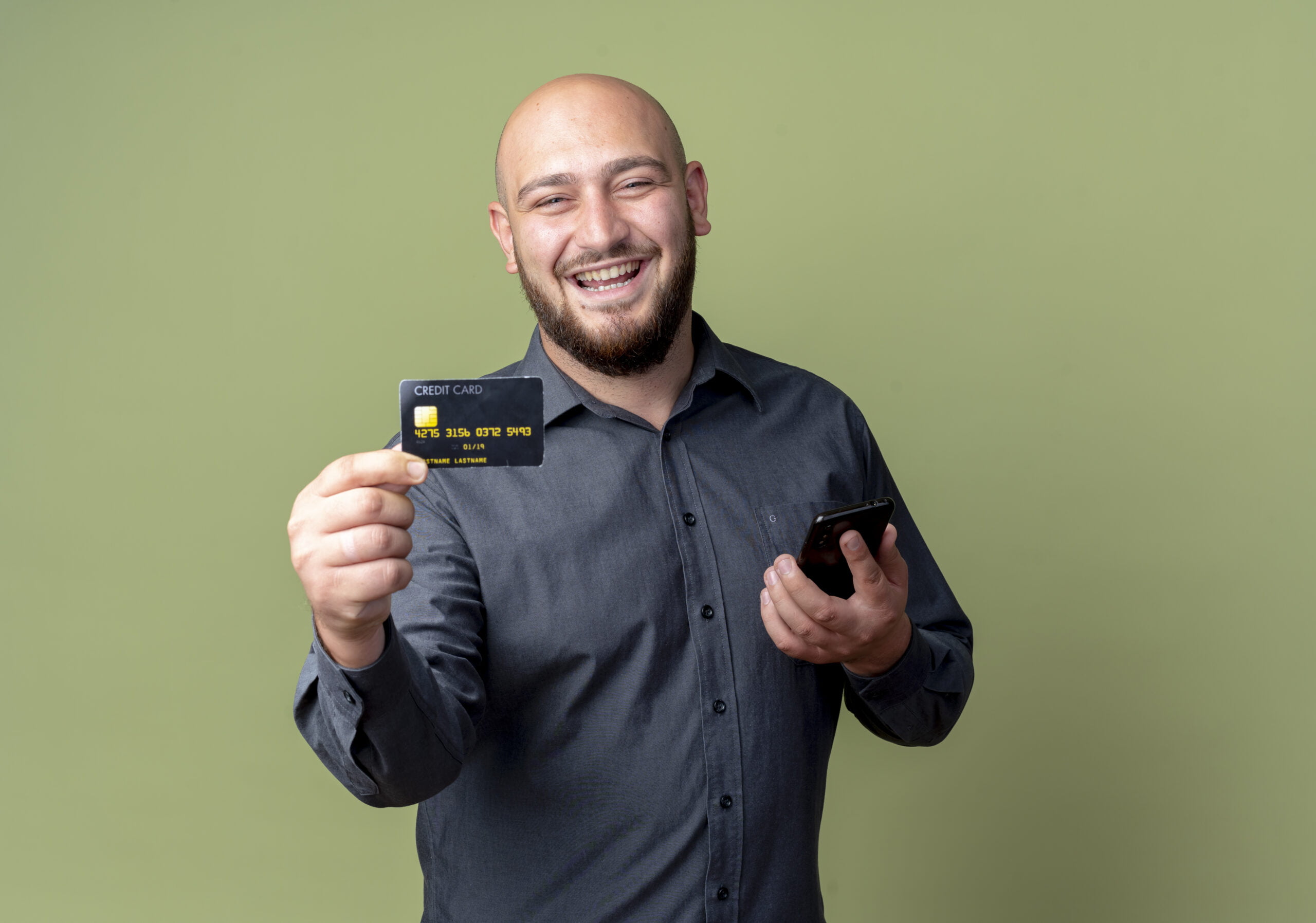 joyful young bald call center man holding mobile phone and stretching out credit card at camera isolated on olive green background with copy space